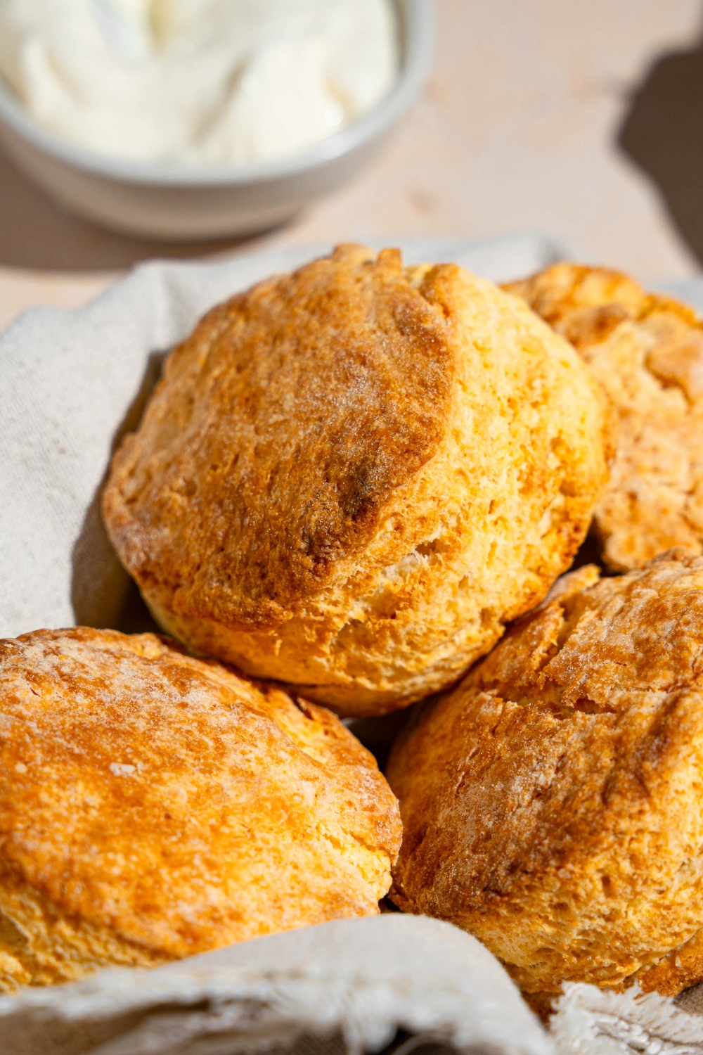 A bowl of shortcake biscuits lined with a cloth napkin. The bowl is on a tan counter with a bowl of whipped cream.