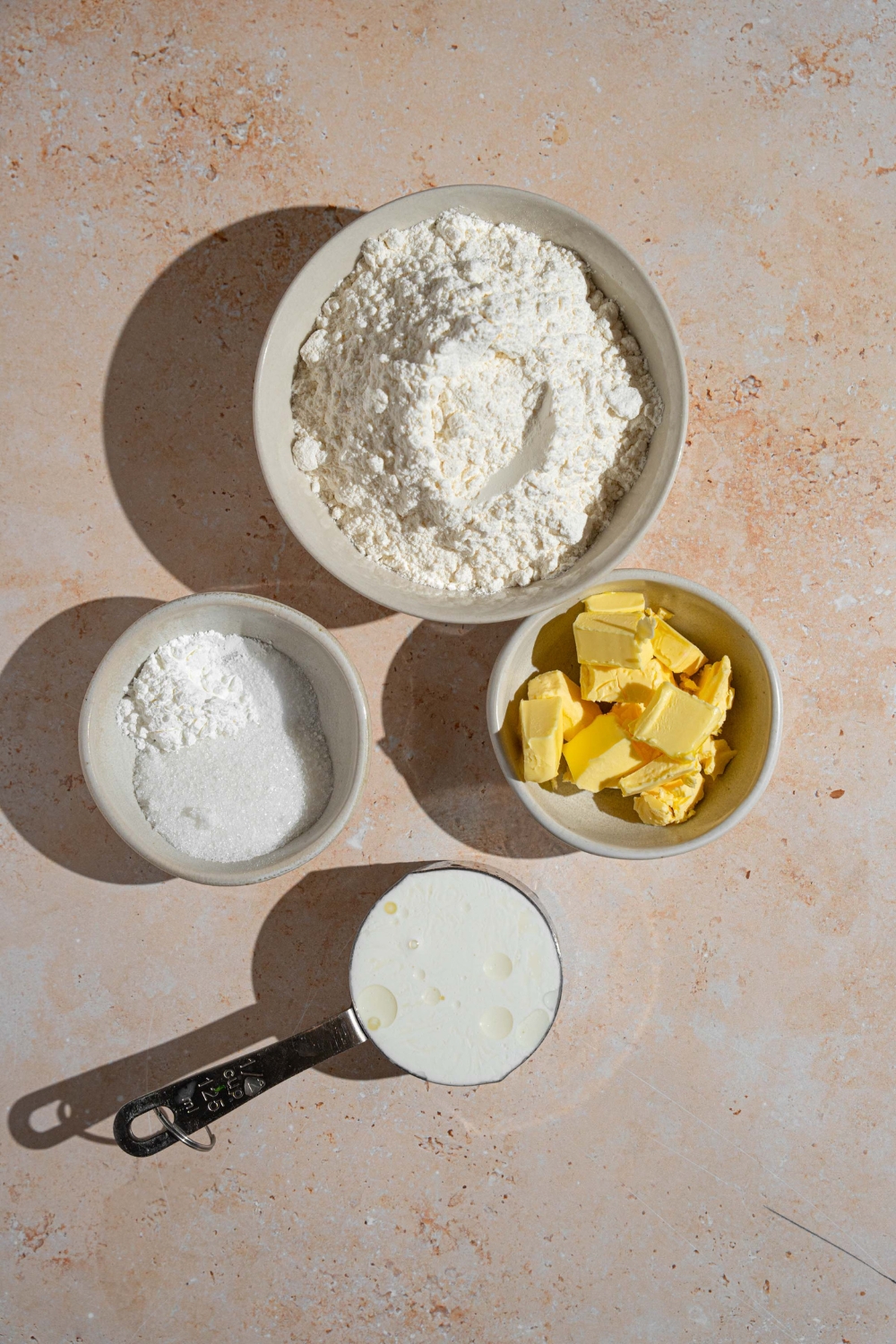 An overhead shot of several bowls in various sizes containing ingredients to make shortcake biscuits including flour, half and half, cold butter, and baking powder.