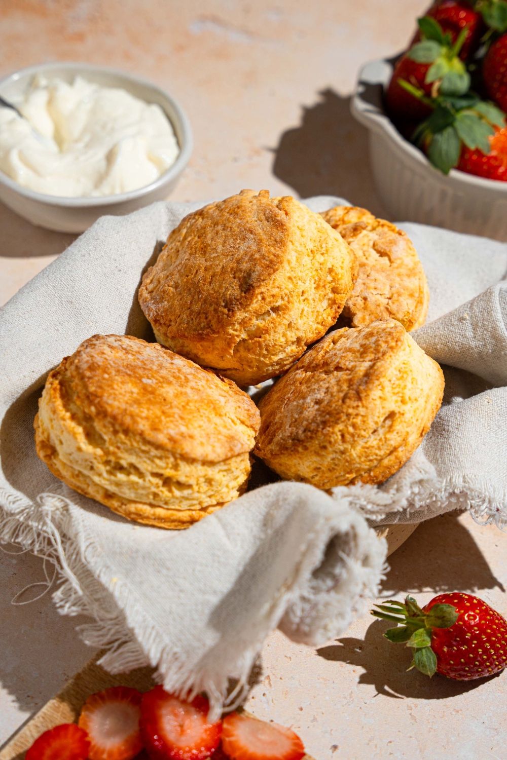 A bowl of shortcake biscuits lined with a cloth napkin. The bowl is on a tan counter with a bowl of whipped cream and bowl of strawberries.