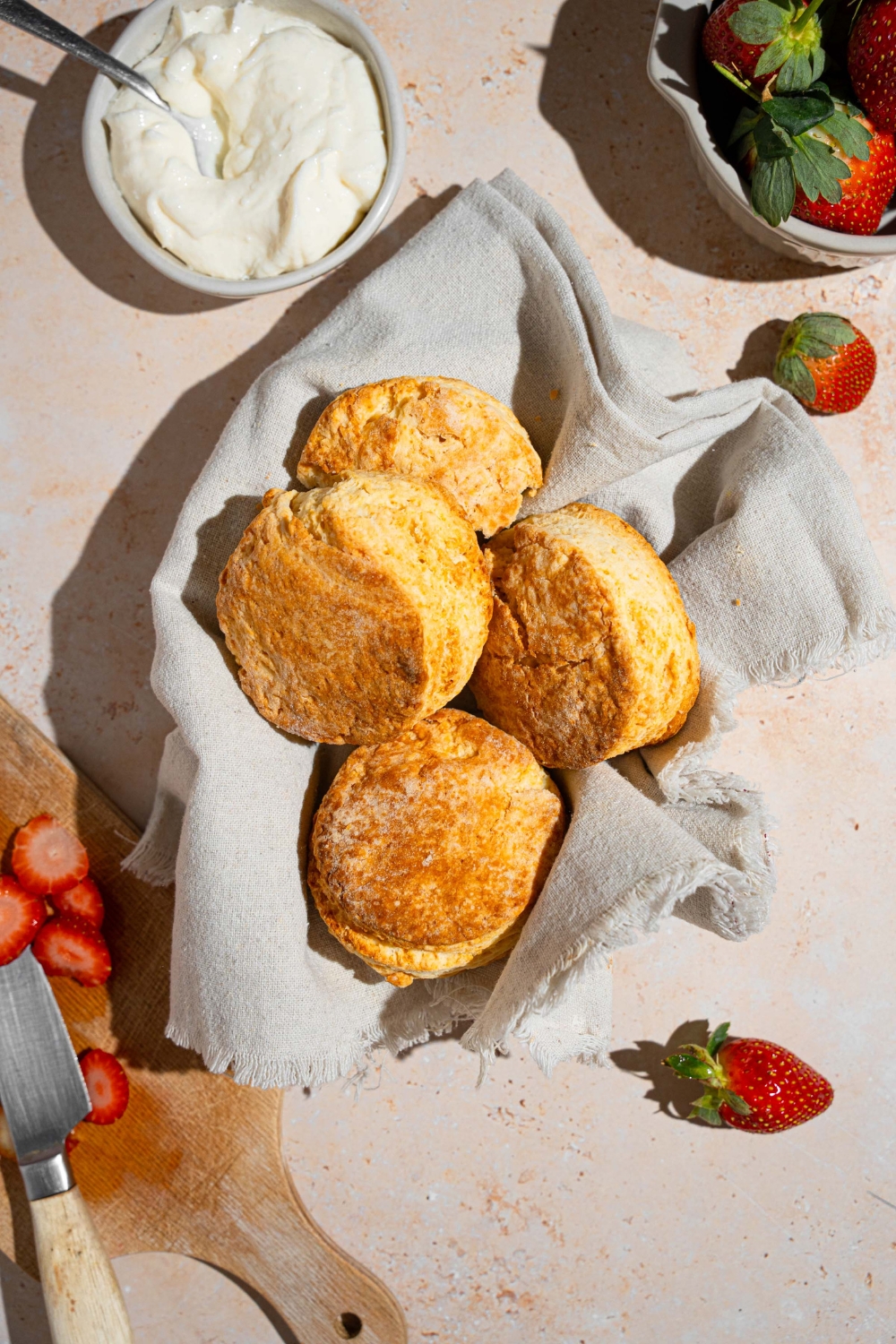 A bowl of shortcake biscuits lined with a cloth napkin. The bowl is on a tan counter with a bowl of whipped cream and bowl of strawberries.