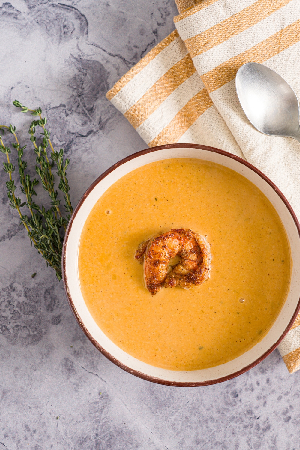 A bowl of shrimp bisque with a piece of shrimp on top. The bowl is on a marble counter with a sprig of thyme, spoon, and yellow checkered napkin.