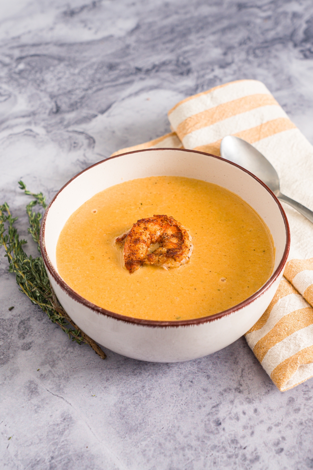 A bowl of shrimp bisque with a piece of shrimp on top. The bowl is on a marble counter with a spoon, sprig of thyme, and yellow checkered napkin.