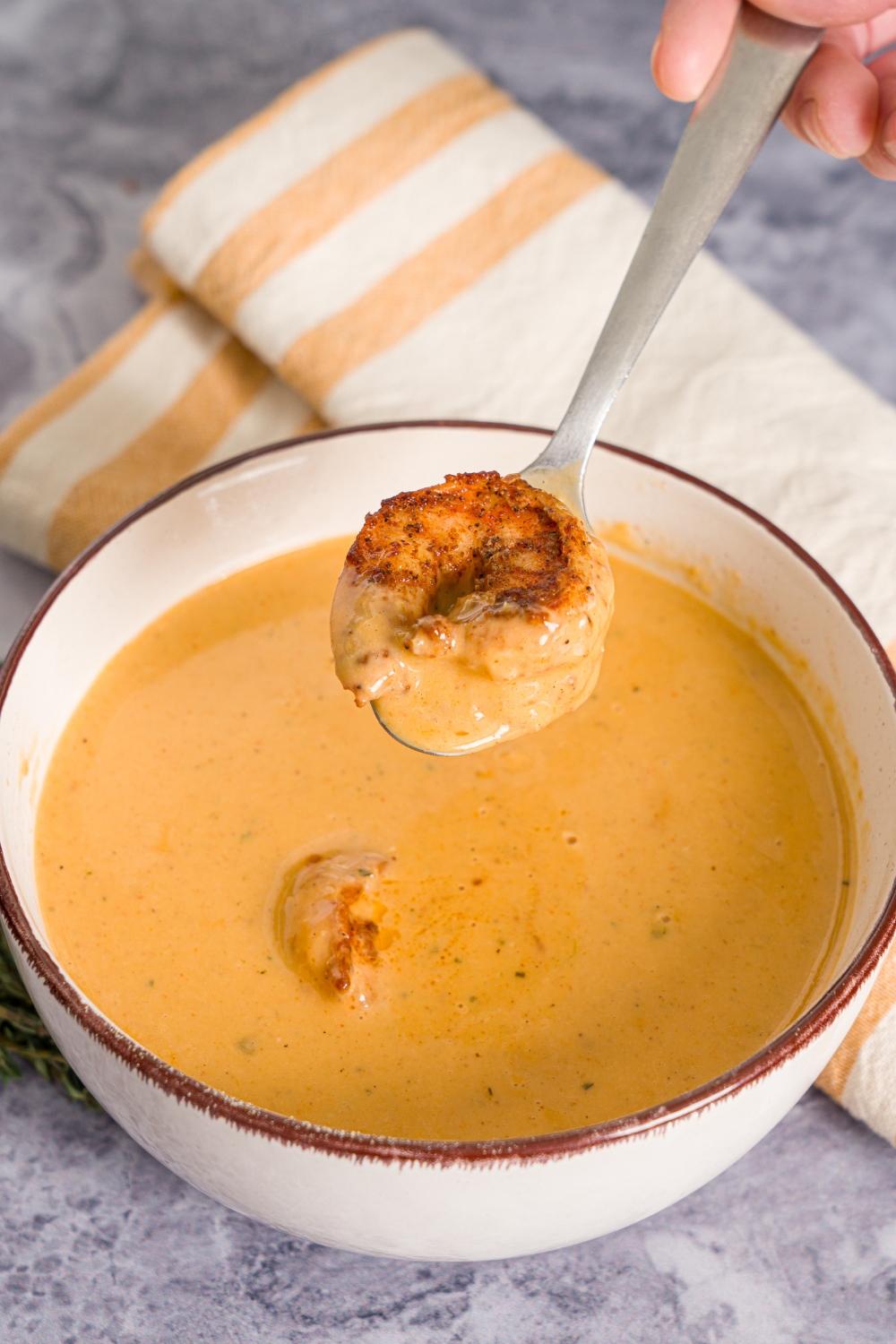 A bowl of shrimp bisque with a spoon dipped in the soup with a shrimp on top. The bowl is on a marble counter with a sprig of thyme, and yellow checkered napkin.