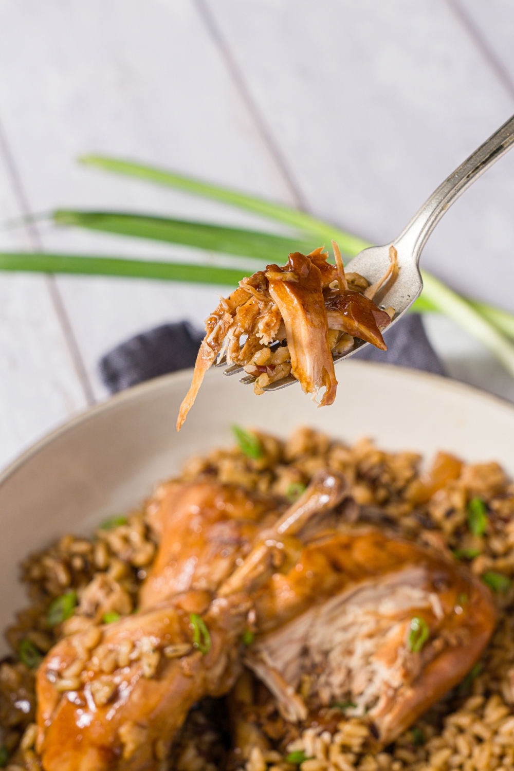 A close up of a fork with a bite of slow cooker chicken adobo. There is a bowl of chicken adobo served over brown rice garnished with sliced green onion blurred in the background.