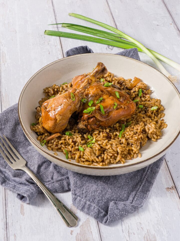 A bowl of slow cooker chicken adobo served over brown rice garnished wtih sliced green onion. The bowl is on a wood counter with a blue cloth napkin and a fork.