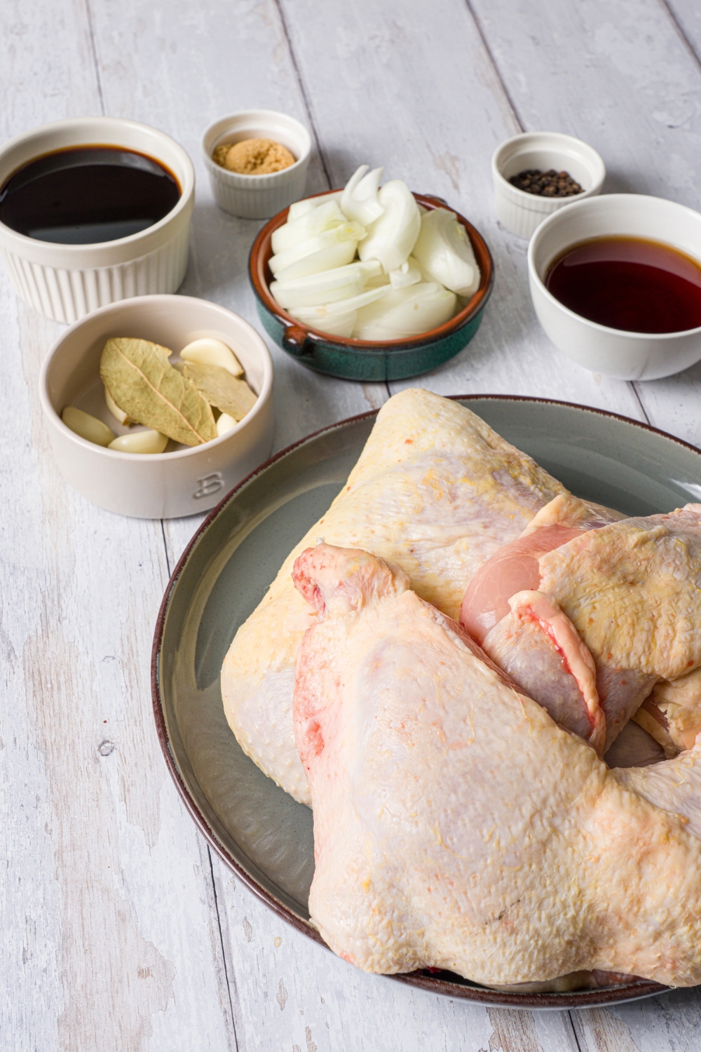 Several bowls in various sizes containing ingredients to make slow cooker chicken adobo including a whole chicken, white onion, soy sauce, red wine vinegar, brown sugar, garlic cloves, and brown sugar.