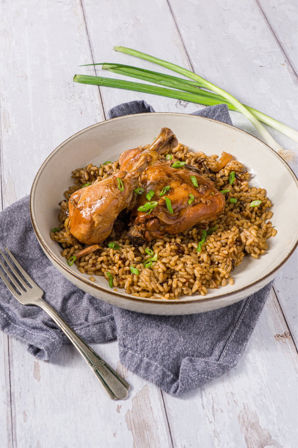 A bowl of slow cooker chicken adobo served over brown rice garnished wtih sliced green onion. The bowl is on a wood counter with a blue cloth napkin and a fork.