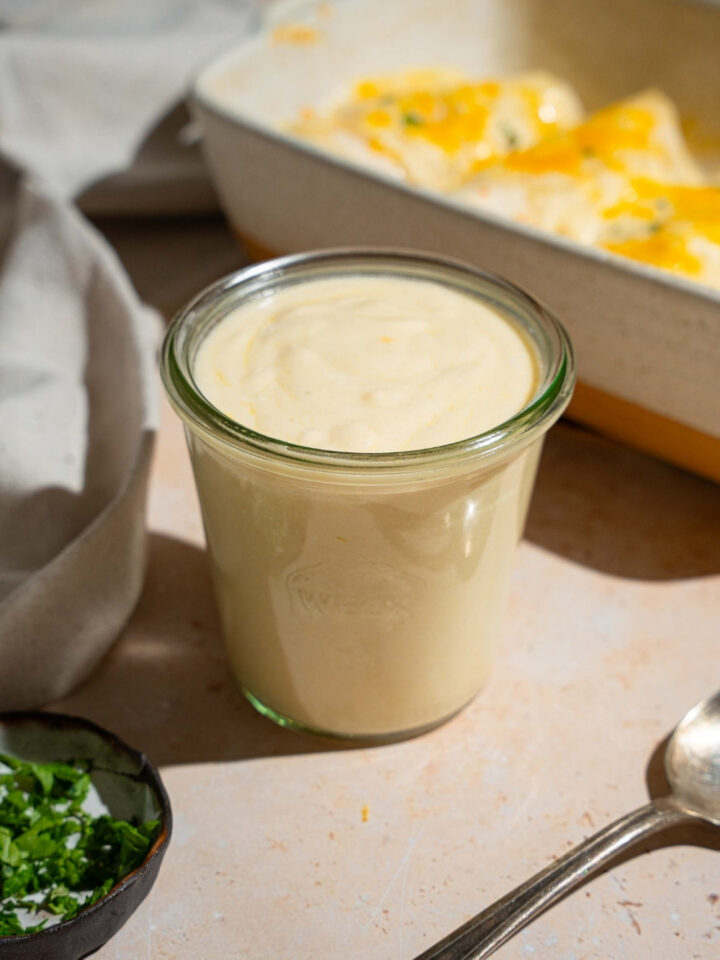 A jar of sour cream sauce for enchiladas. The jar is on a white counter with a baking dish of baked enchiladas and small bowls of garnishes.