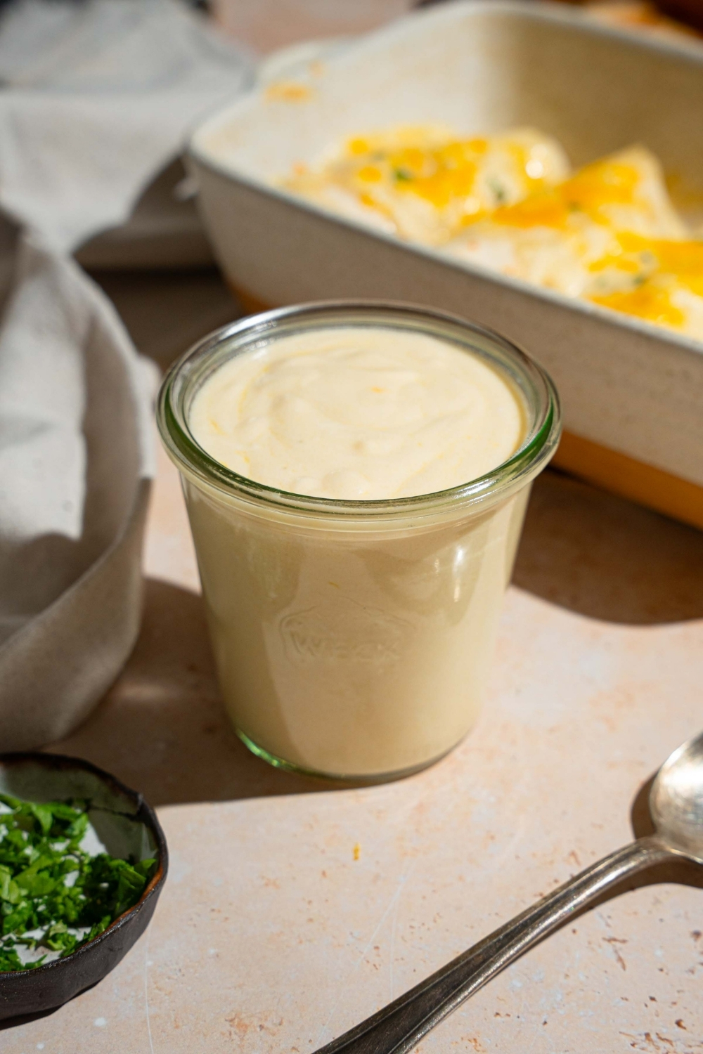 A jar of sour cream sauce for enchiladas. The jar is on a white counter with a baking dish of baked enchiladas and small bowls of garnishes.