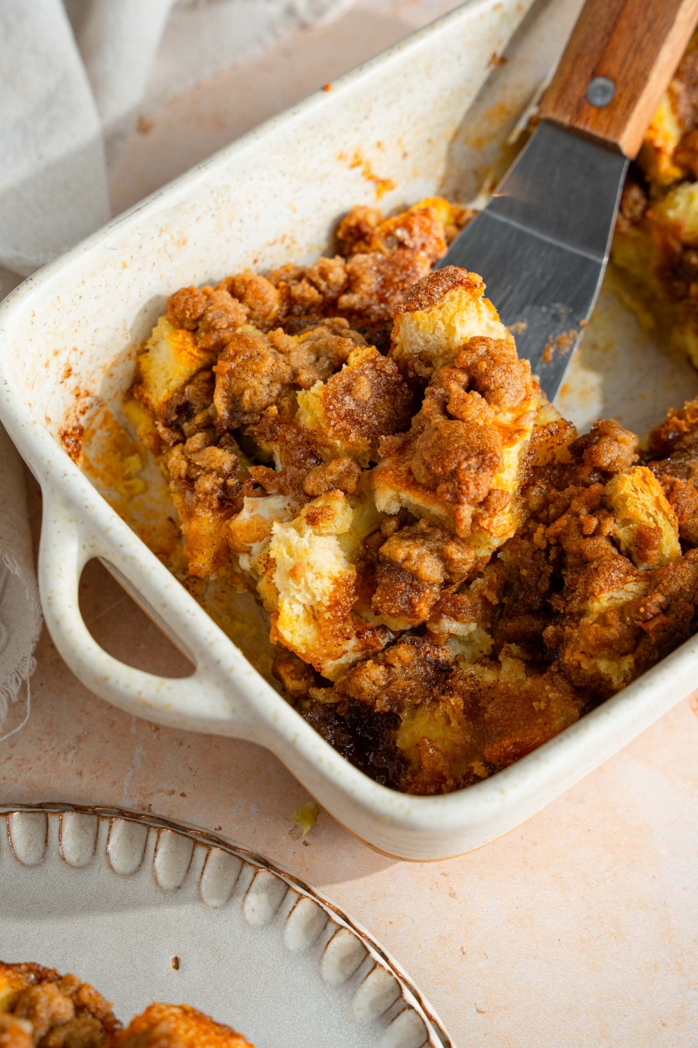 A ceramic baking dish with baked sourdough french toast casserole. There is a spatula removing a slice of french toast from the dish. The dish is on a tan counter with a white cloth napkin.