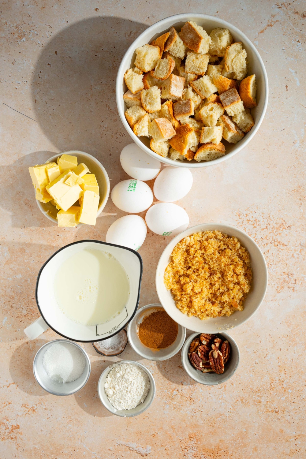 An overhead shot of several bowls in various sizes containing ingredients to make sourdough french toast casserole including sliced sourdough bread, butter, eggs, brown sugar, cinnamon, heavy cream, flour, and pecans.