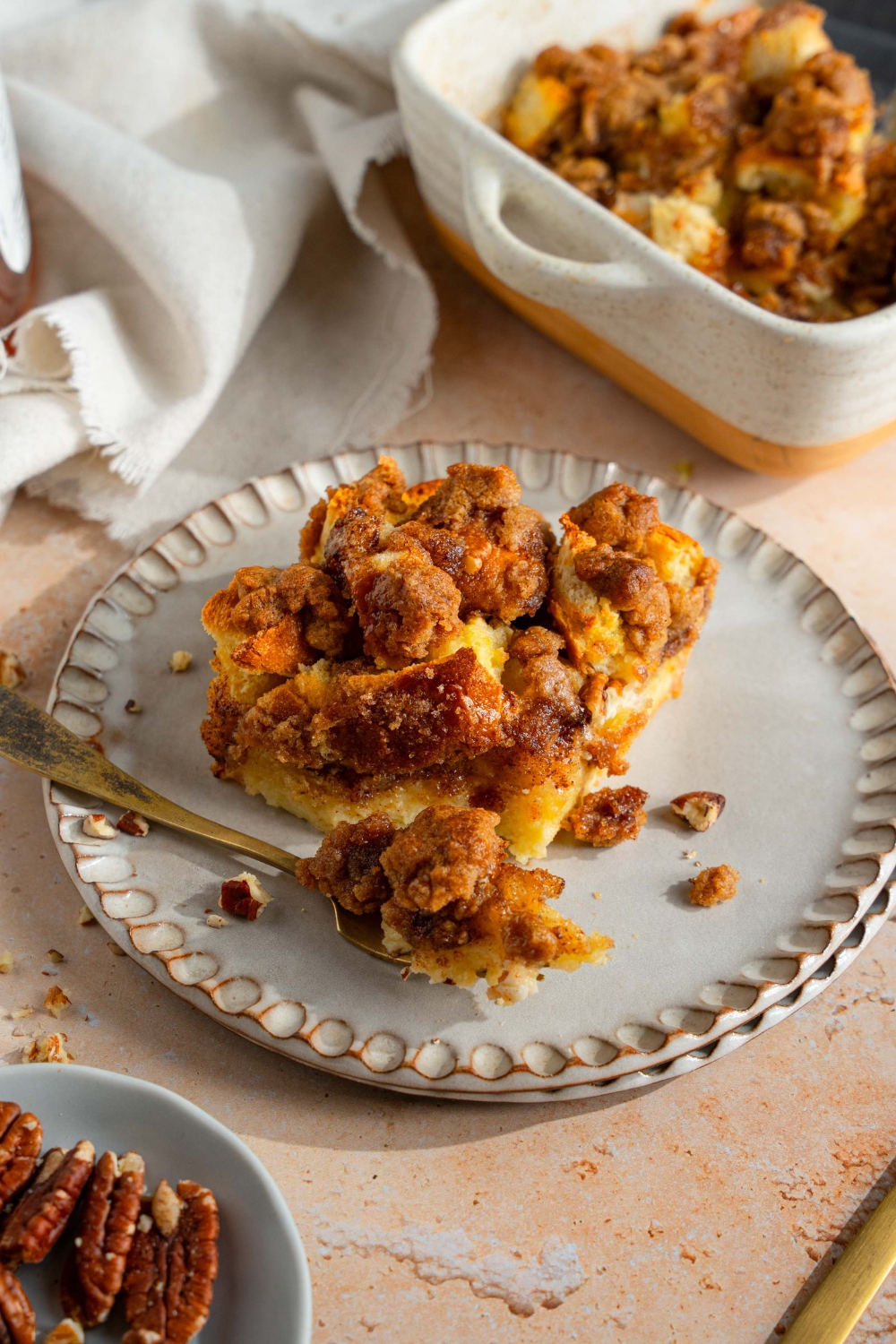 A white plate with a slice of sourdough french toast casserole. There is a fork with a bite of french toast on the plate. The plate is on a tan counter with a baking dish of french toast and white cloth napkin.