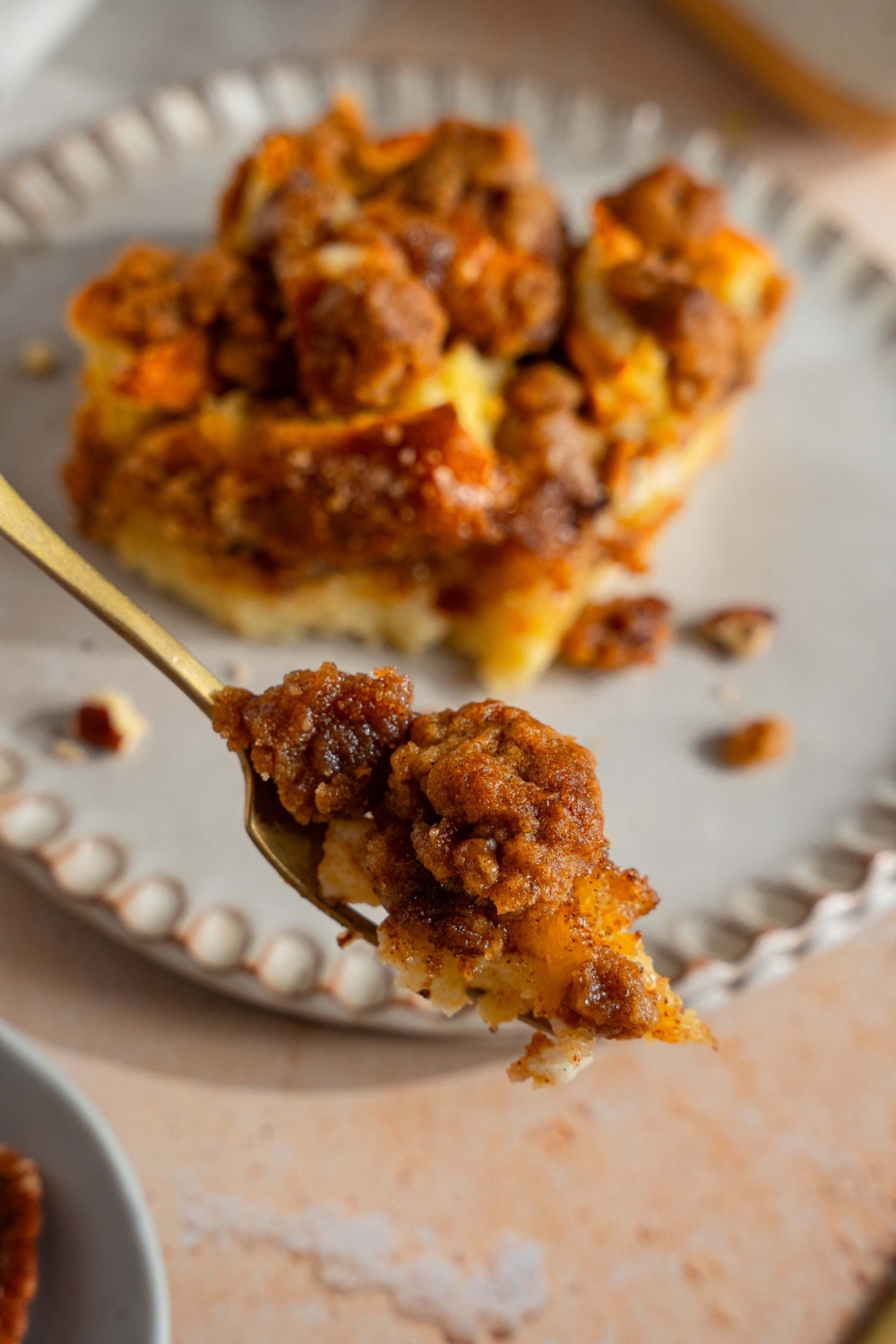 A close up of a fork with a bite of sourdough french toast casserole with a plate of french toast blurred in the background.