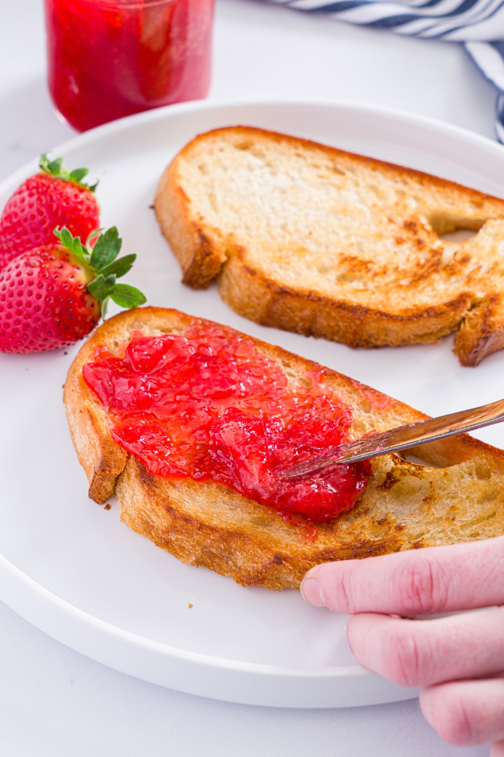 A white plate with two slices of toast with a knife spreading strawberry jam on the toast. The plate is on a marble counter with a jar of strawberry jam.