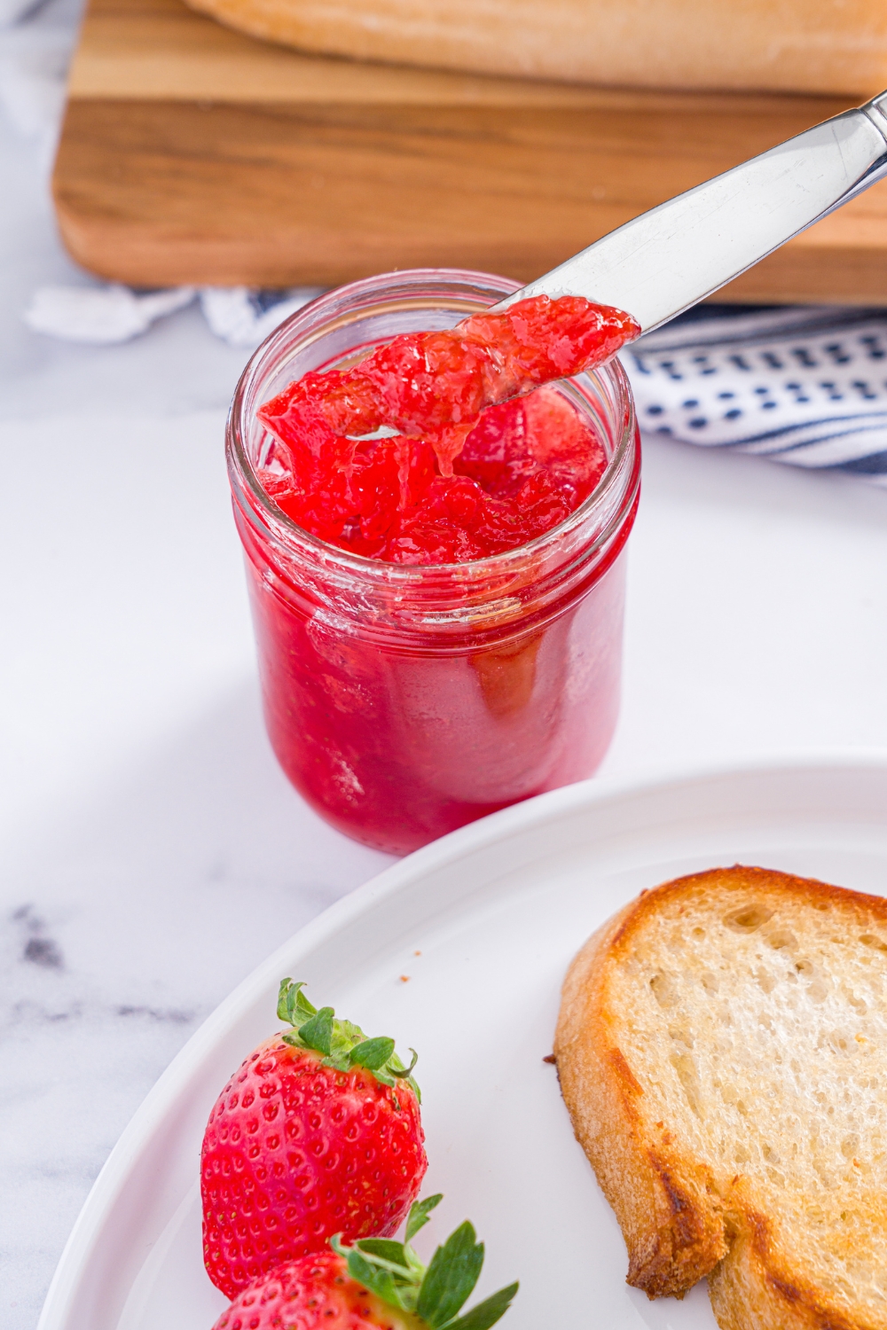A jar of strawberry jam with a knife dipped into the jar. The jar is on a marble counter with a plate of toast and strawberries.