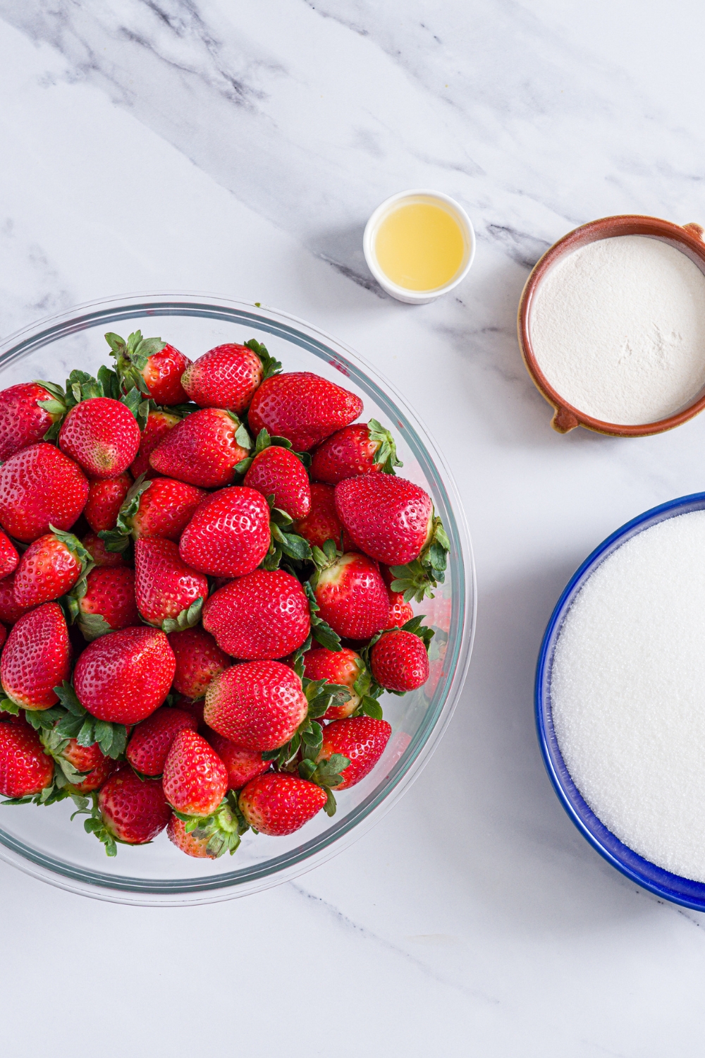 A marble counter with bowls of ingredients to make strawberry jam including a bowl of strawberries, sugar, pectin, and lemon juice.