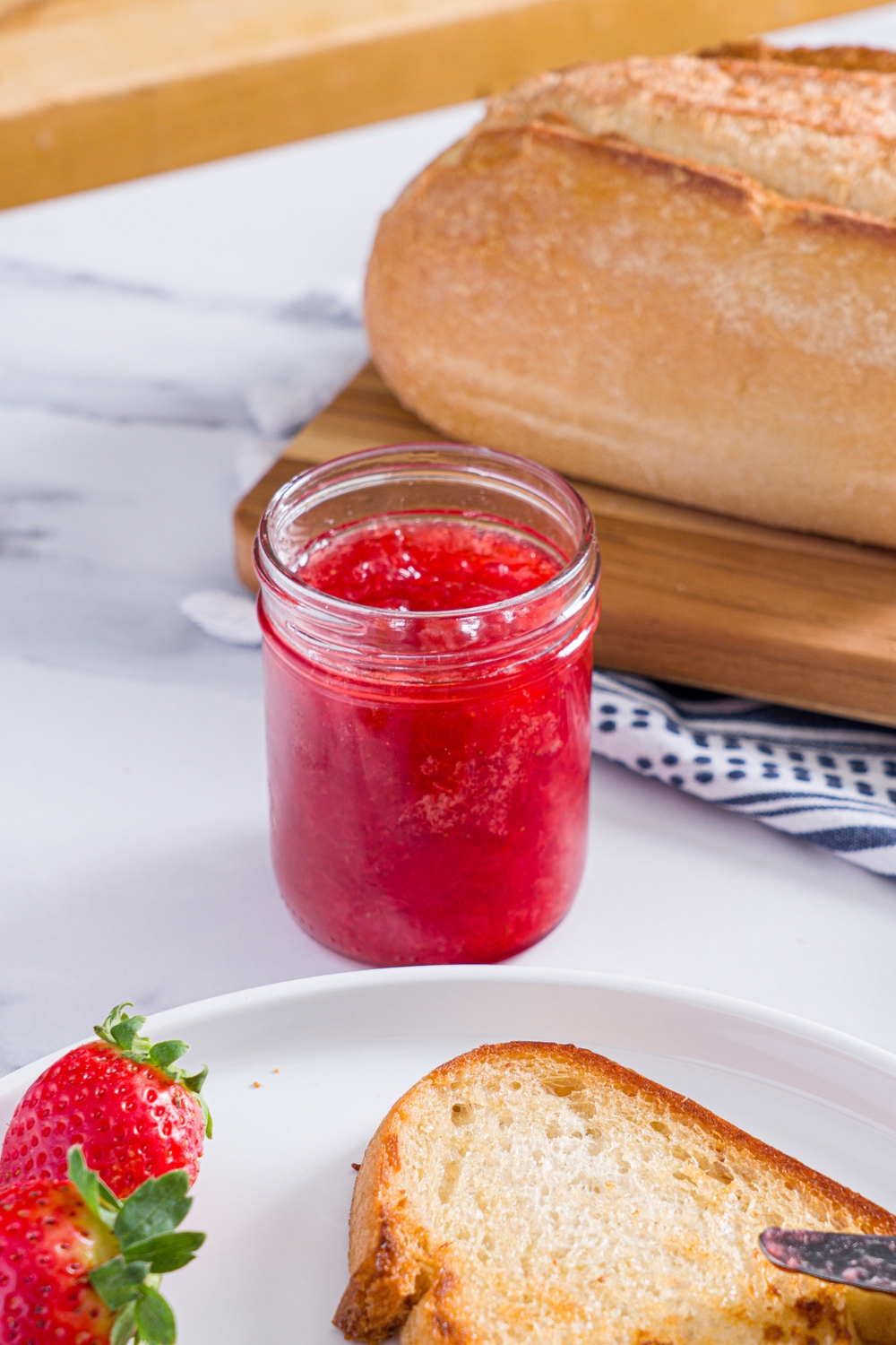 A jar of strawberry jam. The jar is on a marble counter with a plate of toast and strawberries and a board of bread.