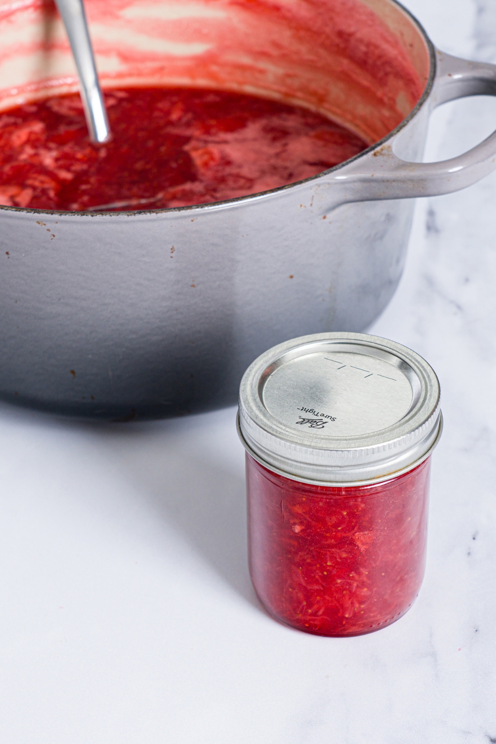 A canned and sealed jar of strawberry jam next to a dutch oven with cooked strawberry jam. The pot is on a marble counter.