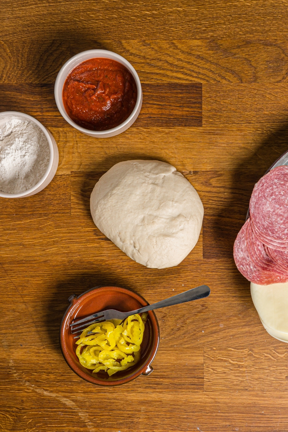 A wood counter with ingredients to make stromboli including a ball of pizza dough, bowl of pizza sauce, bowl of sliced banana peppers, and bowl of flour.