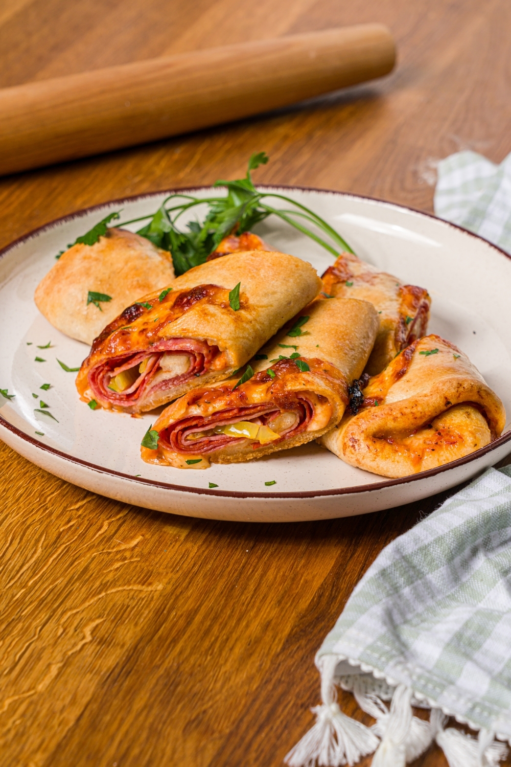 A white plate with sliced stromboli garnished with fresh parsley. The plate is on a wood counter with a wooden rolling pin and green checkered napkin.