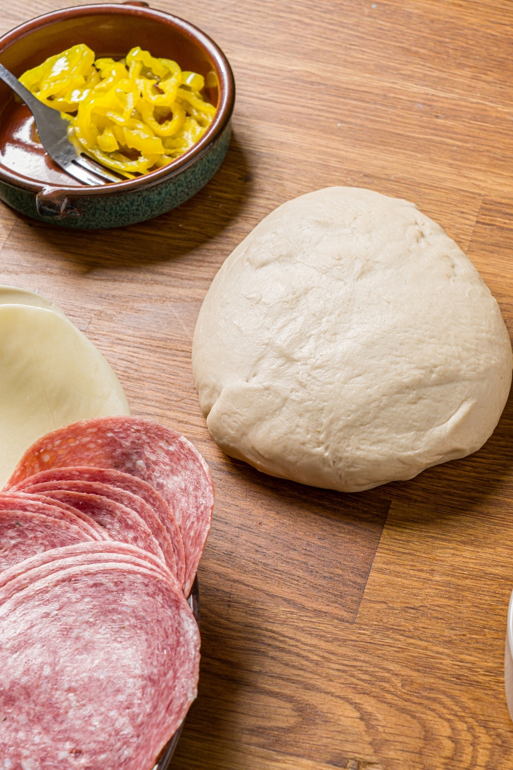 A wood counter with a ball of pizza dough, small bowl of sliced banna peppers, and plate of sliced meats and cheeses.