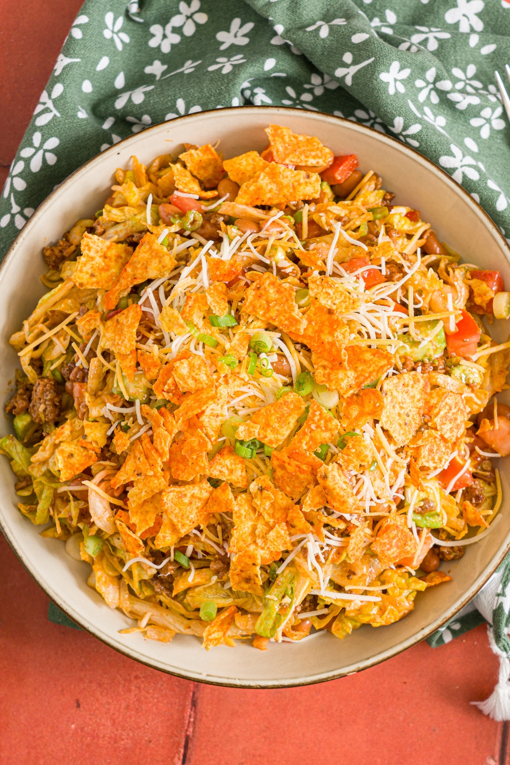 A white bowl with taco salad tossed with shredded lettuce, ground taco meat, shredded cheese, diced tomatoes, diced avocados, and green onion tossed with catalina dressing and topped with doritos. The bowl is on a red tiled counter with a green floral napkin.