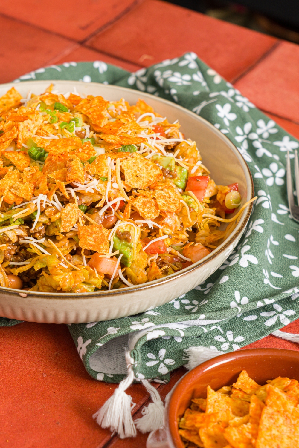 A white bowl with taco salad tossed with shredded lettuce, ground taco meat, shredded cheese, diced tomatoes, diced avocados, and green onion tossed with catalina dressing and topped with doritos. The bowl is on a red tiled counter with a green floral napkin.