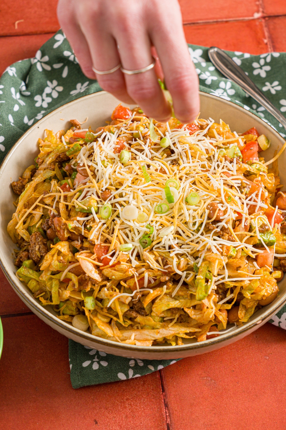 A white bowl of taco salad with shredded lettuce, cooked taco meat, diced tomato, and shredded cheese. A hand is sprinkling sliced green onions over the bowl. The bowl is on a red tiled counter with a green floral napkin.