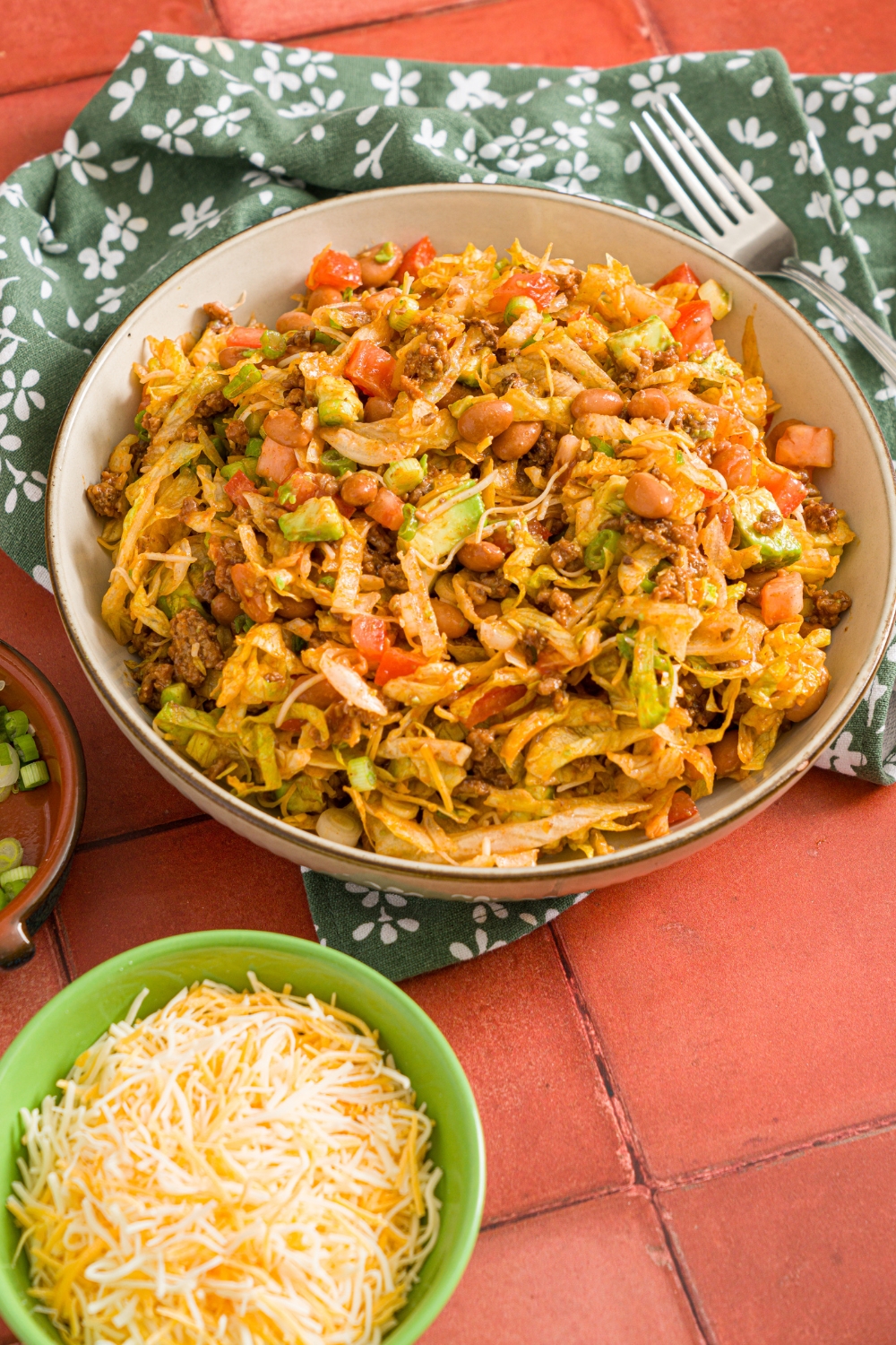 A white bowl with taco salad including shredded lettuce, taco meat, chili beans, diced tomatoes, shredded cheese, and diced avocados tossed in catalina dressing. The bowl is on a red tiled counter with a green floral napkin and small bowl of shredded cheese.