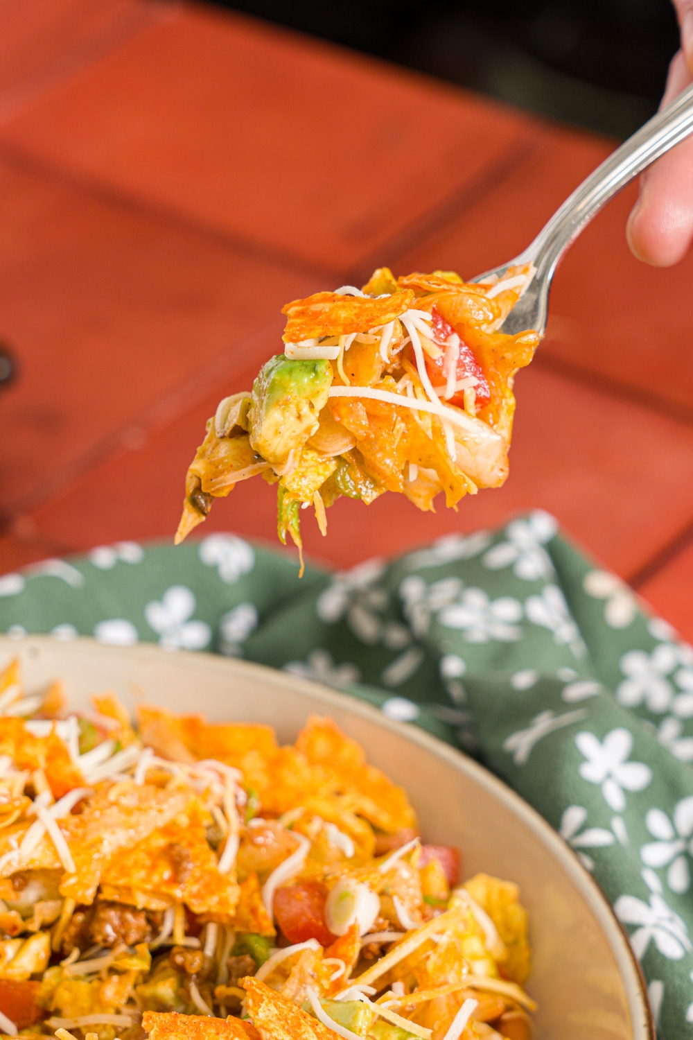 A fork with a bite of taco salad with catalina dressing, doritos, avocado, and shredded cheese. The fork is over a bowl of taco salad on a red tiled counter with a green floral napkin.