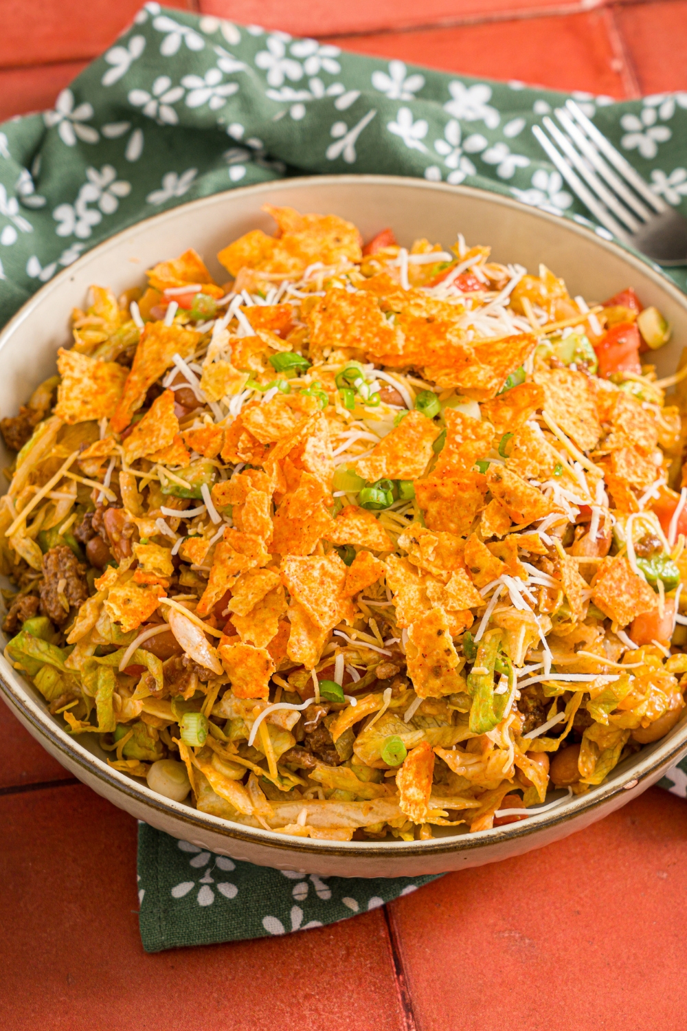 A white bowl with taco salad tossed with shredded lettuce, ground taco meat, shredded cheese, diced tomatoes, diced avocados, and green onion tossed with catalina dressing and topped with doritos. The bowl is on a red tiled counter with a green floral napkin.