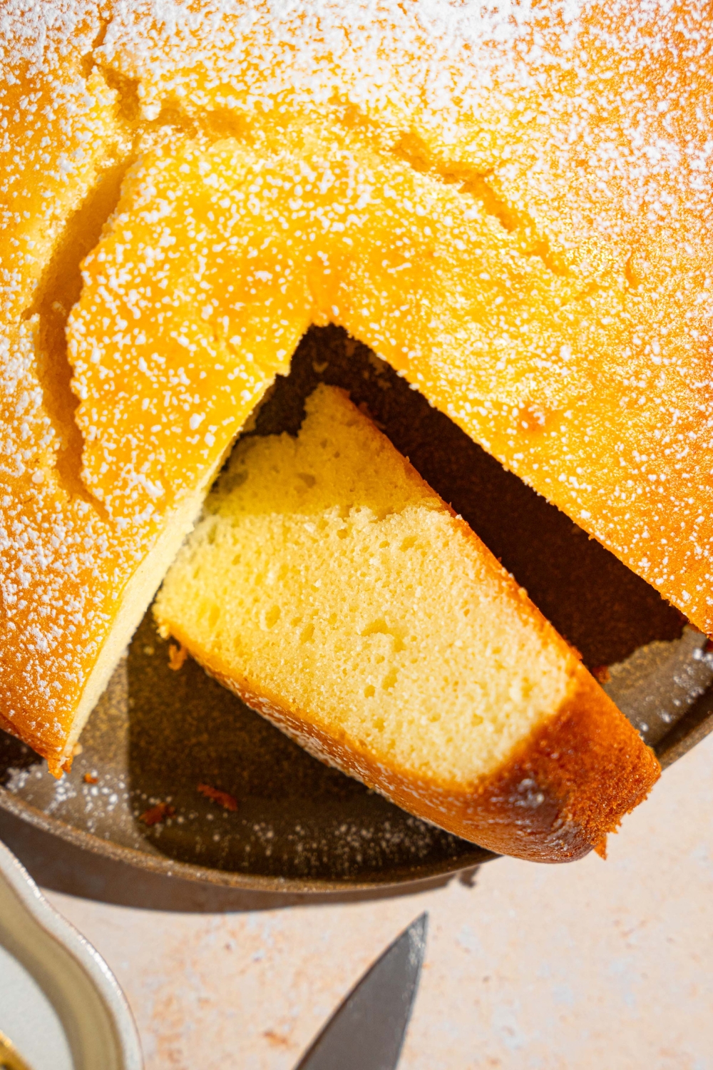 A green plate with a whole French butter cake dusted with powdered sugar. There is a slice removed from the cake on the plate. The plate is on a tan counter.