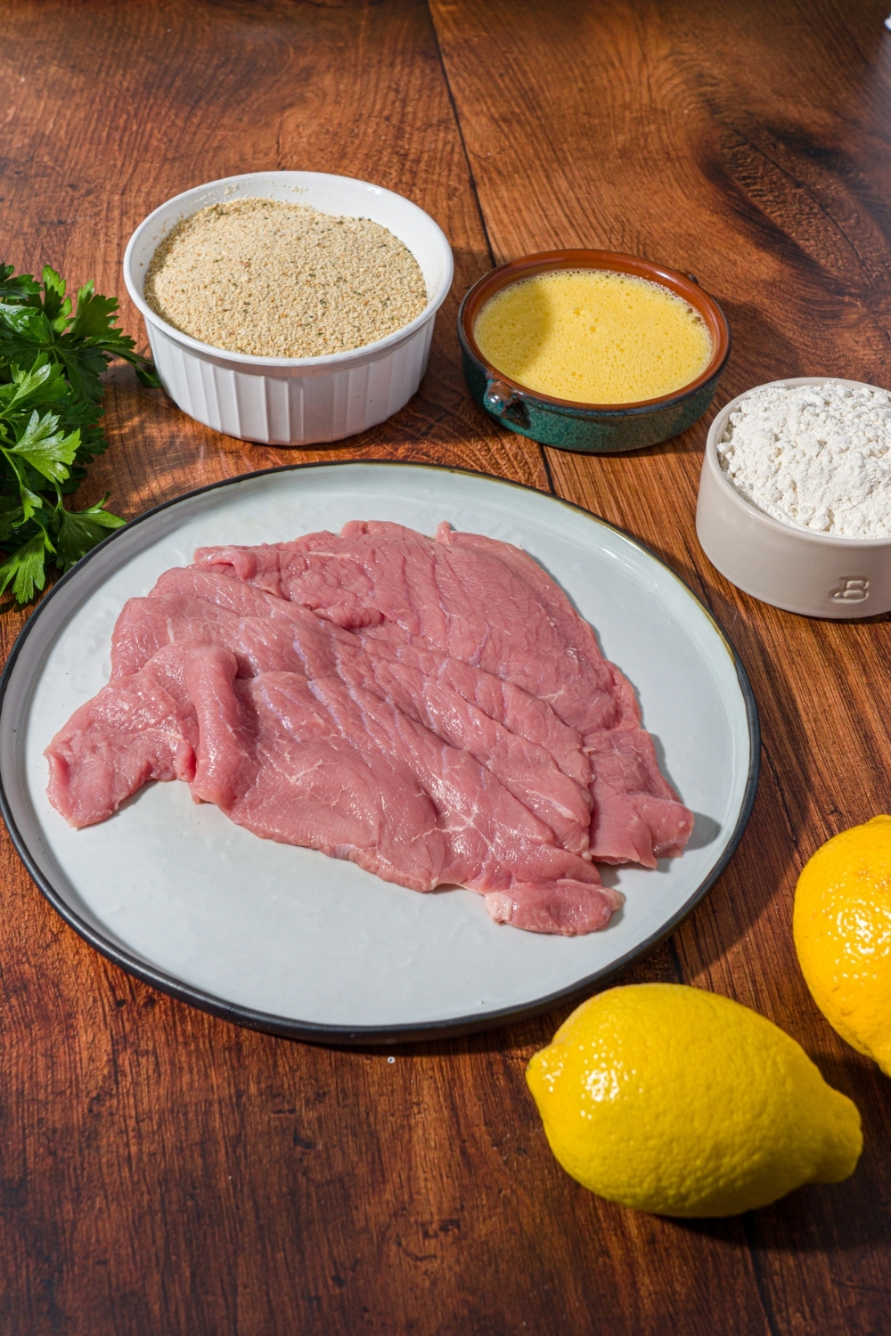 A wood counter with ingredients to make veal cutlets including veal, breadcrumbs, egg, flour, fresh parsley, and lemon