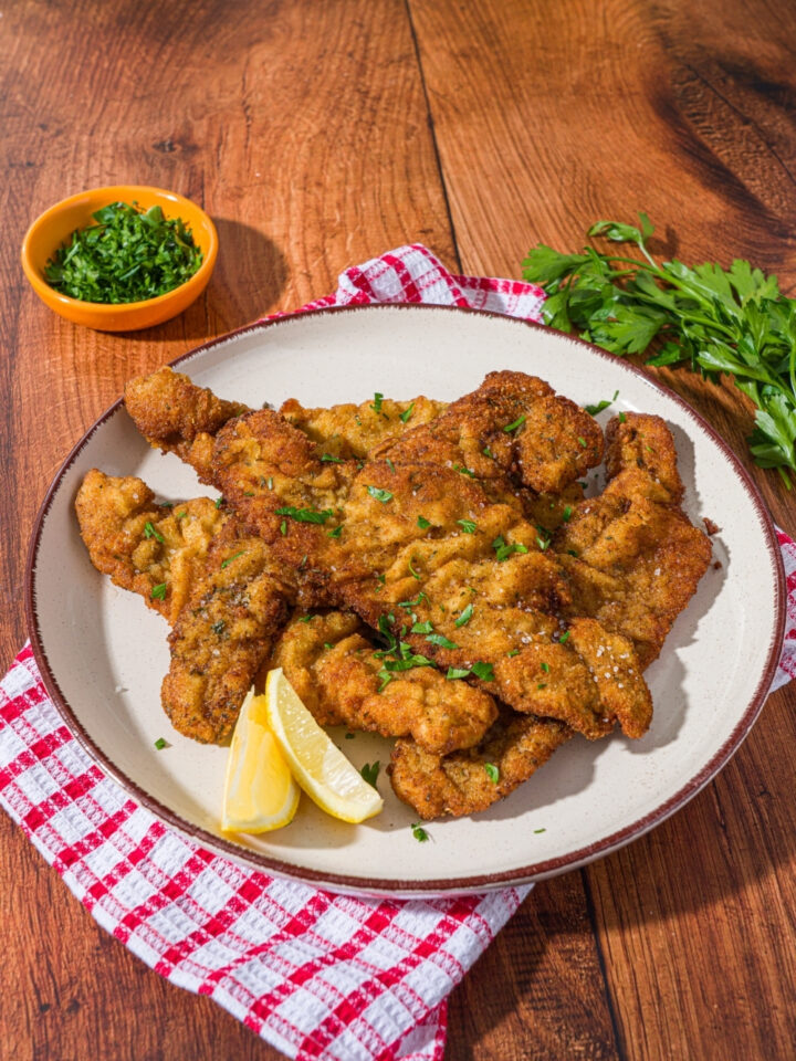 A white plate with breaded veal cutlets garnished with fresh parsley and served with lemon wedges. The plate is on a red checkered napkin on a wood counter with a small bowl of garnishes.