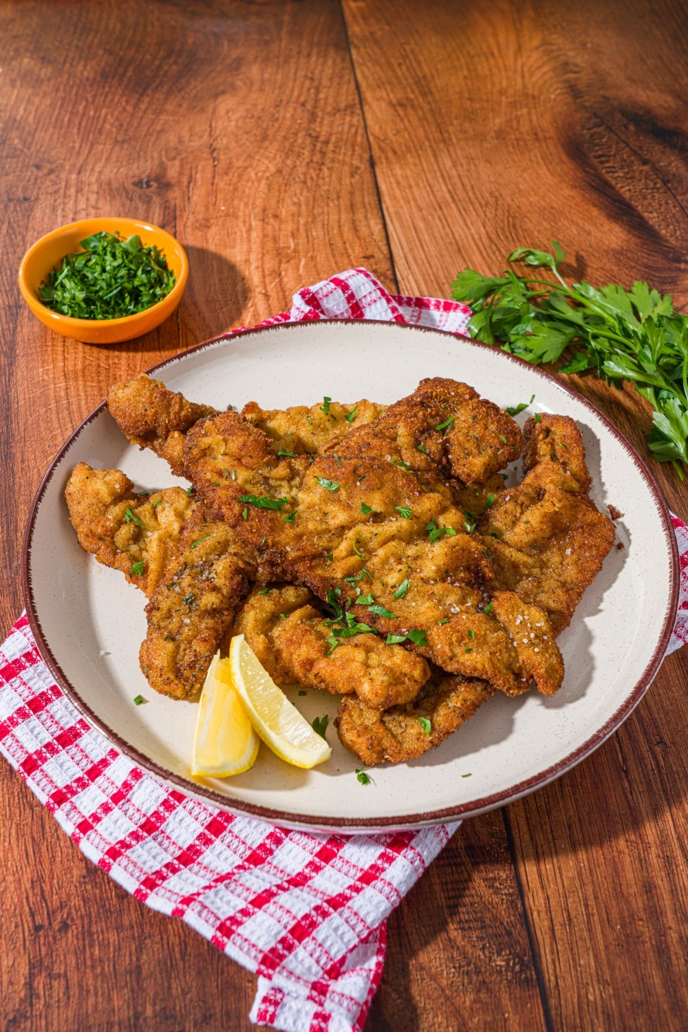 A white plate with breaded veal cutlets garnished with fresh parsley and served with lemon wedges. The plate is on a red checkered napkin on a wood counter with a small bowl of garnishes.
