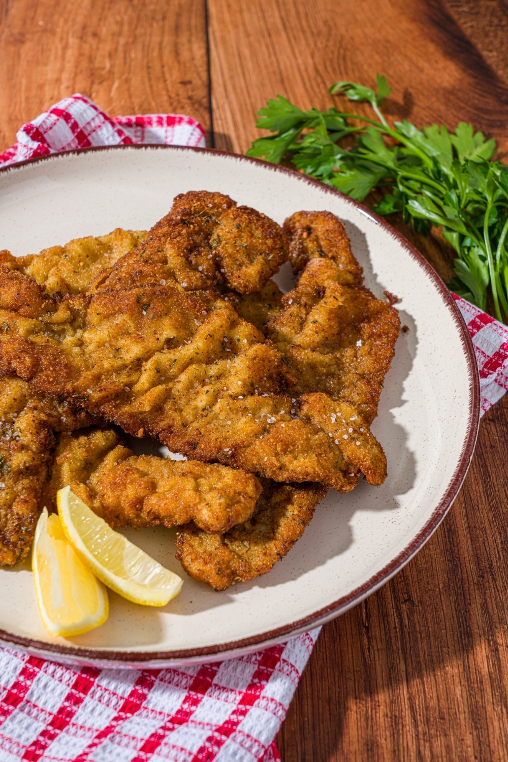 A white plate with breaded veal cutlets served with lemon wedges. The plate is on a red checkered napkin on a wood counter with a small bowl of garnishes.