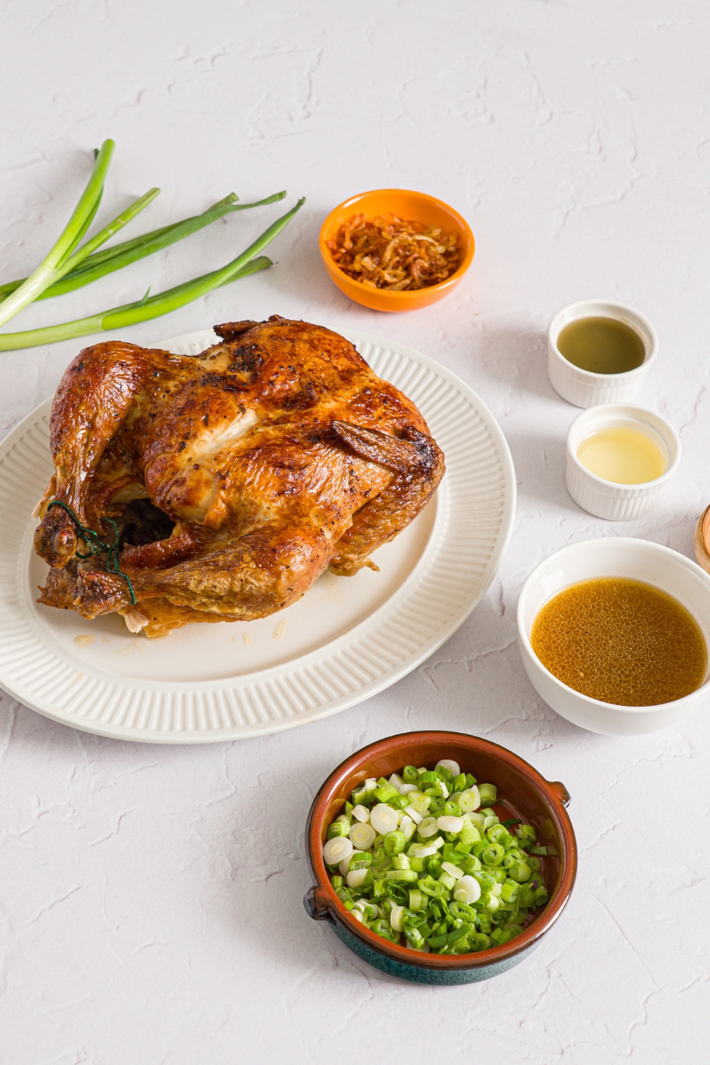 A white counter with several bowls in various sizes containing ingredients to make viral Costco chicken, scallion, and rice including a whole rotisserie chicken, sliced scallions, chicken juices, golden shallot oil, scallion sauce, and crispy fried onions.