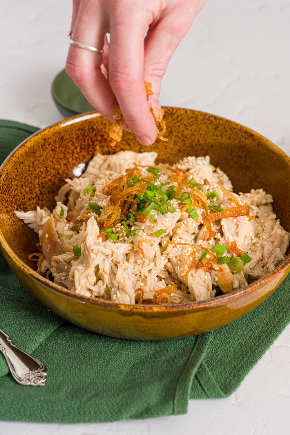 A brown ceramic bowl with viral Costco chicken, scallion, and rice topped with sliced scallions. A hand is sprinkling crispy fried onions over the bowl. The bowl is on a white counter with a green cloth napkin.