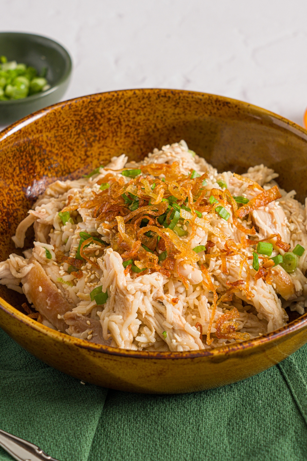 A brown ceramic bowl with viral Costco chicken, scallion, and rice topped with sliced scallions and crispy fried onions. The bowl is on a white counter with a green cloth napkin and small bowl of sliced scallions.