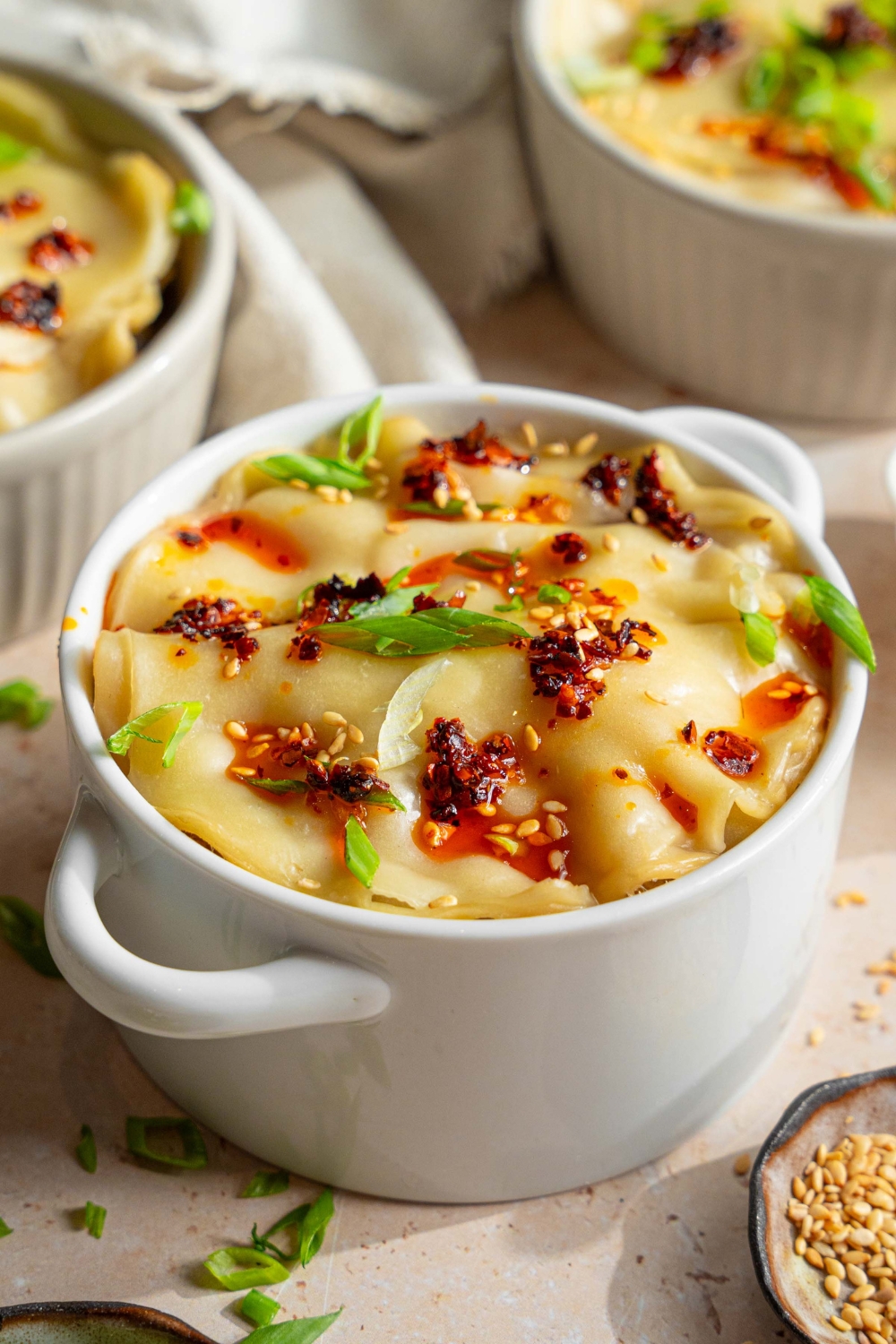 A ramekin with dumpling lasagna topped with chili oil, sliced green onions, and sesame seeds. The ramekin is on a tan counter with additional ramekins of dumpling lasagna, a white cloth napkin, and a small bowls of garnishes.