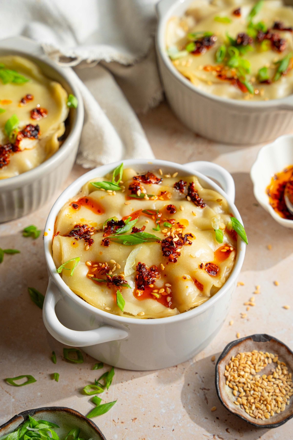 A ramekin with dumpling lasagna topped with chili oil, sliced green onions, and sesame seeds. The ramekin is on a tan counter with additional ramekins of dumpling lasagna, a white cloth napkin, and a small bowls of garnishes.