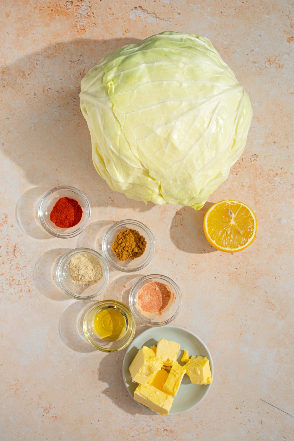 An overhead shot of several bowls in various sizes containing ingredients to make cabbage boil including a whole head of cabbage, butter, lemon, smoked paprika, cajun seasoning, Old Bay seasoning, and olive oil.