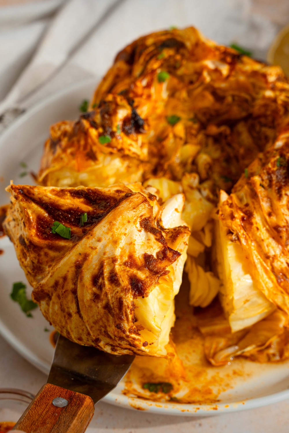 A plate with a baked cabbage boil with cajun seasoned butter. There is a spatula taking a piece of cabbage from the plate. The plate is on a tan counter with a small bowl of seasoned butter and fresh parsley.
