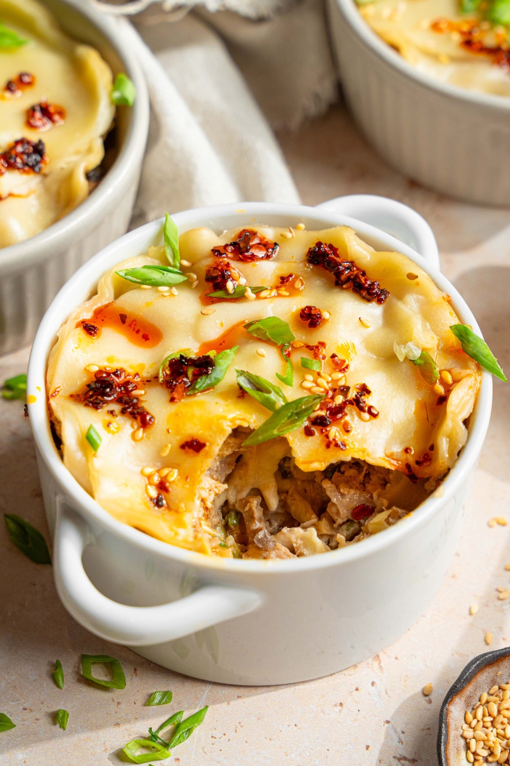 A ramekin with dumpling lasagna topped with chili oil, sliced green onions, and sesame seeds. A bite is taken from the lasagna. The ramekin is on a tan counter with additional ramekins of dumpling lasagna, a white cloth napkin, and a small bowls of garnishes.
