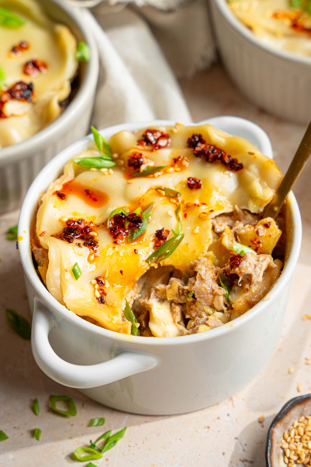 A ramekin with dumpling lasagna topped with chili oil, sliced green onions, and sesame seeds. A fork is taking a bite of lasagna. The ramekin is on a tan counter with additional ramekins of dumpling lasagna, a white cloth napkin, and a small bowls of garnishes.