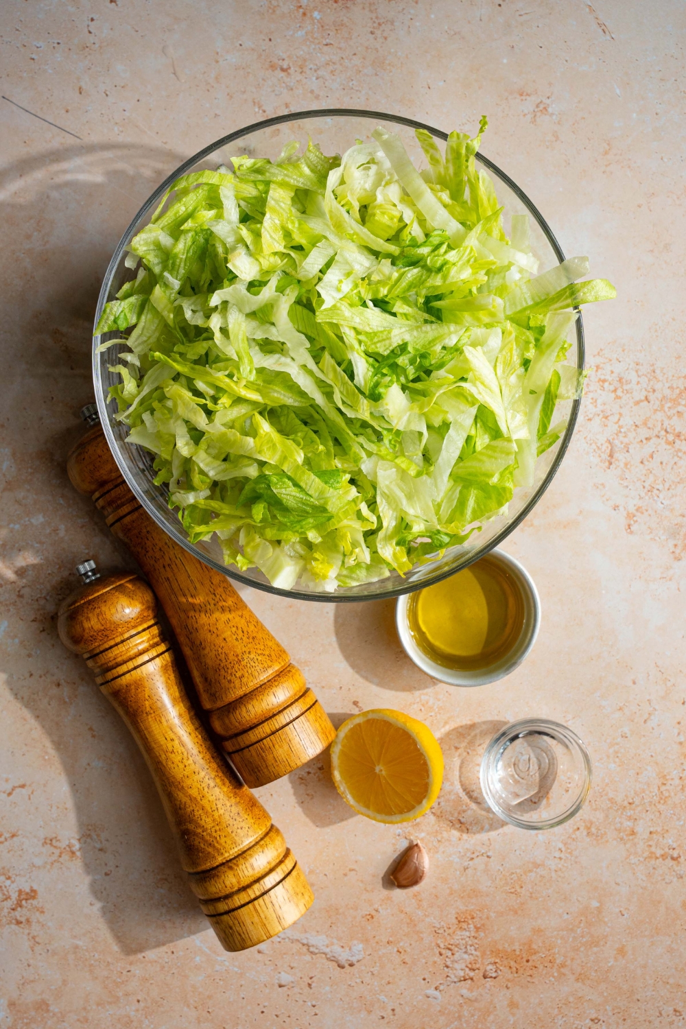An overhead shot of several ingredients to make iceberg lettuce salad including a glass bowl with chopped iceberg lettuce, a bowl of oil, half of a lemon, a bowl of vinegar, garlic, and a salt and pepper mill.