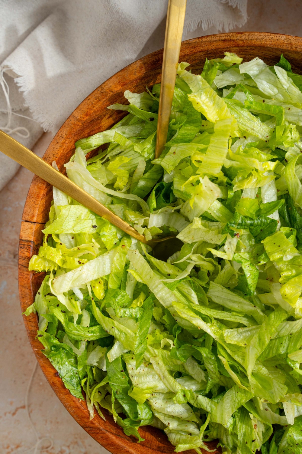 A wooden salad bowl with iceberg lettuce salad tossed in lemon dressing. There are two forks in the salad. The bowl is on a tan counter with a white cloth napkin.