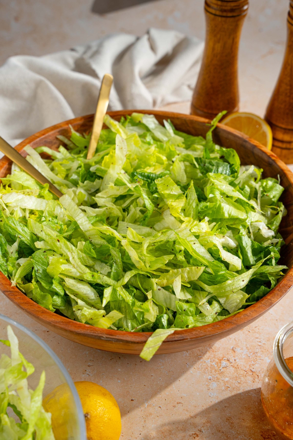 A wooden salad bowl with iceberg lettuce salad tossed in lemon dressing. There are two forks in the salad. The bowl is on a tan counter with a salt and pepper mill, lemons, and a white cloth napkin.