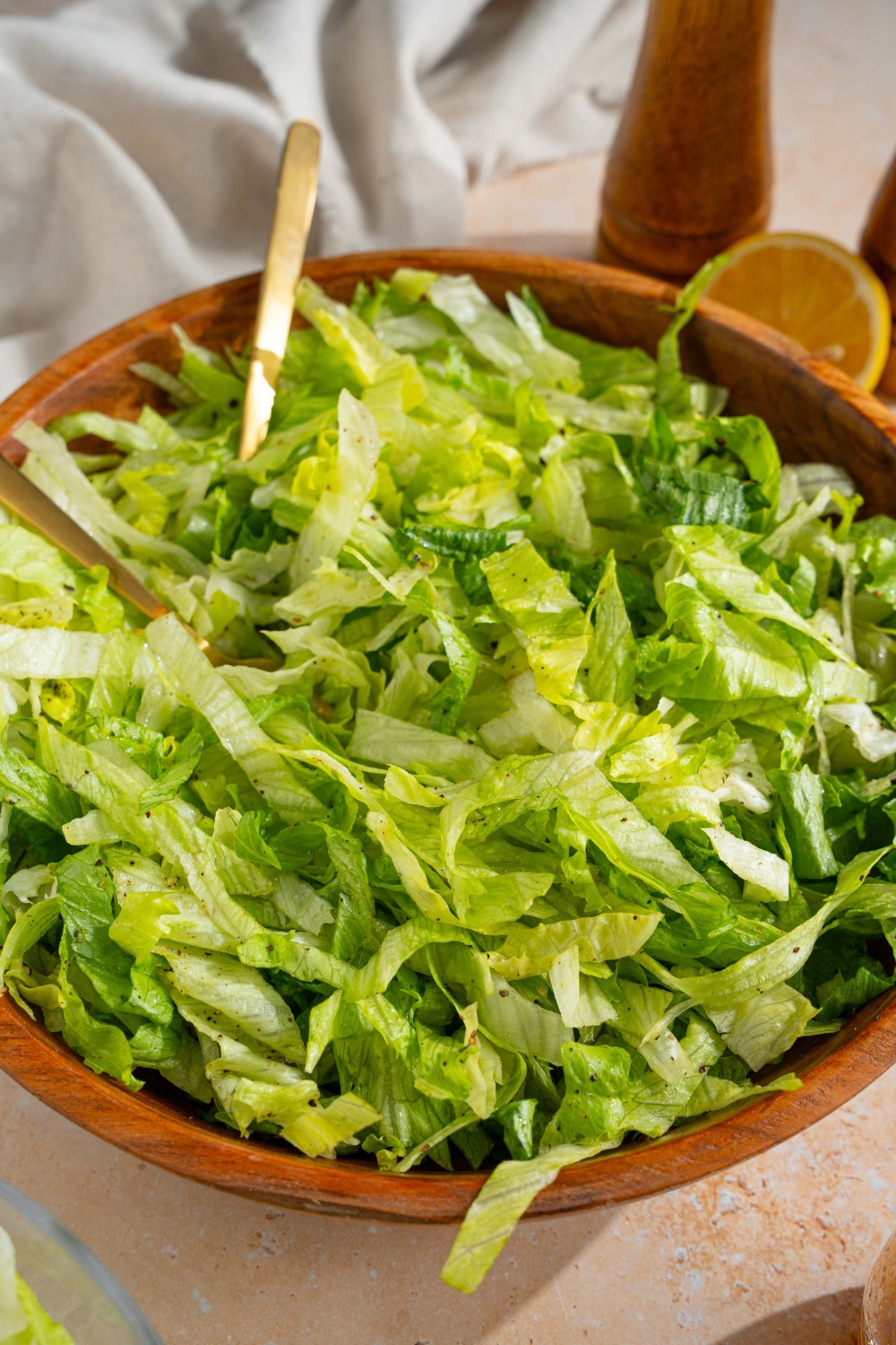 A wooden salad bowl with iceberg lettuce salad tossed in lemon dressing. There are two forks in the salad. The bowl is on a tan counter with a salt and pepper mill, lemons, and a white cloth napkin.
