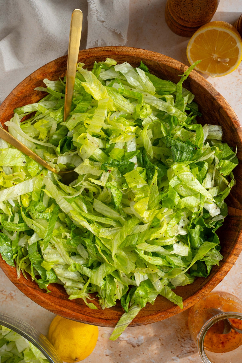 A wooden salad bowl with iceberg lettuce salad tossed in lemon dressing. There are two forks in the salad. The bowl is on a tan counter with a salt and pepper mill, lemons, and a white cloth napkin.